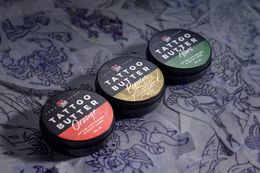 Three tins of Love Ink Tattoo Butter arranged in a row on a background of tattoo sketches. From left to right: 'Tattoo Butter Orange' with an orange design, 'Tattoo Butter Papaya' with a yellow design, and 'Tattoo Butter Aloe' with a green design. All tins are 100 ml and are intended for after tattooing and daily care.