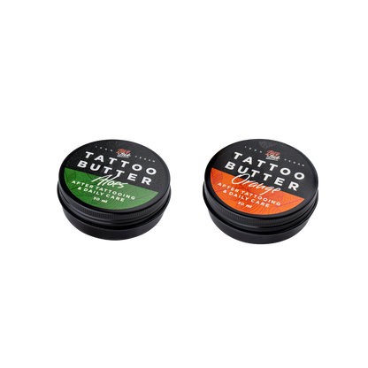 Two tins of Love Ink Tattoo Butter on a transparent background. One tin is labeled 'Tattoo Butter Aloes' with an green design, and the other is labeled 'Tattoo Butter Orange' with an orange design. Both tins are 50 ml and are intended for after tattooing and daily care.