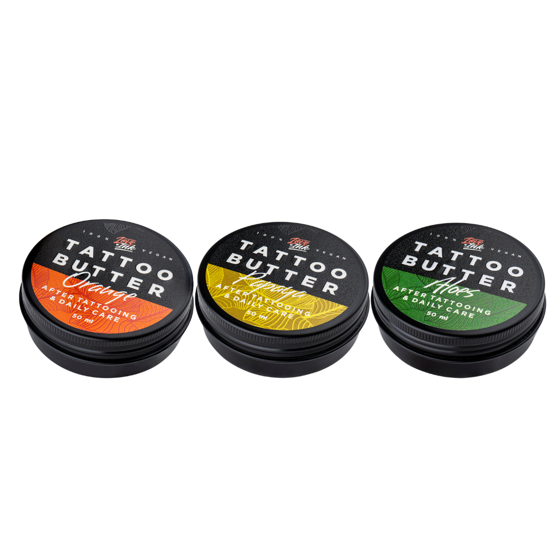 Three tins of Love Ink Tattoo Butter on a transparent background. From left to right: 'Tattoo Butter Orange' with an orange design, 'Tattoo Butter Papaya' with a yellow design, and 'Tattoo Butter Aloe' with a green design. All tins are 50 ml and are intended for after tattooing and daily care.