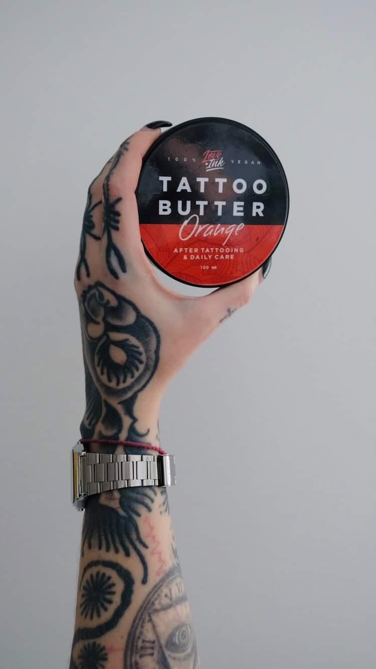 Tattooed hand displaying a jar of Tattoo Butter Orange by LoveInk against a plain background. The product is 100% vegan and intended for after tattooing and daily care, 100ml.
