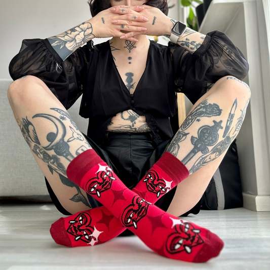 Tattooed person sitting on the floor with legs crossed, wearing red socks with a stylized heart design featuring horns and fangs, in a brightly lit indoor setting.