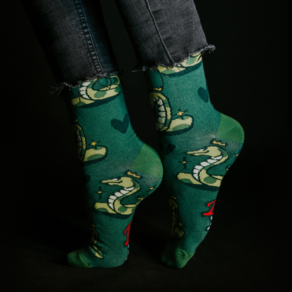 Close-up of feet wearing green LOVEINK socks with a snake and crown design, paired with frayed-edge black jeans, set against a dark background.