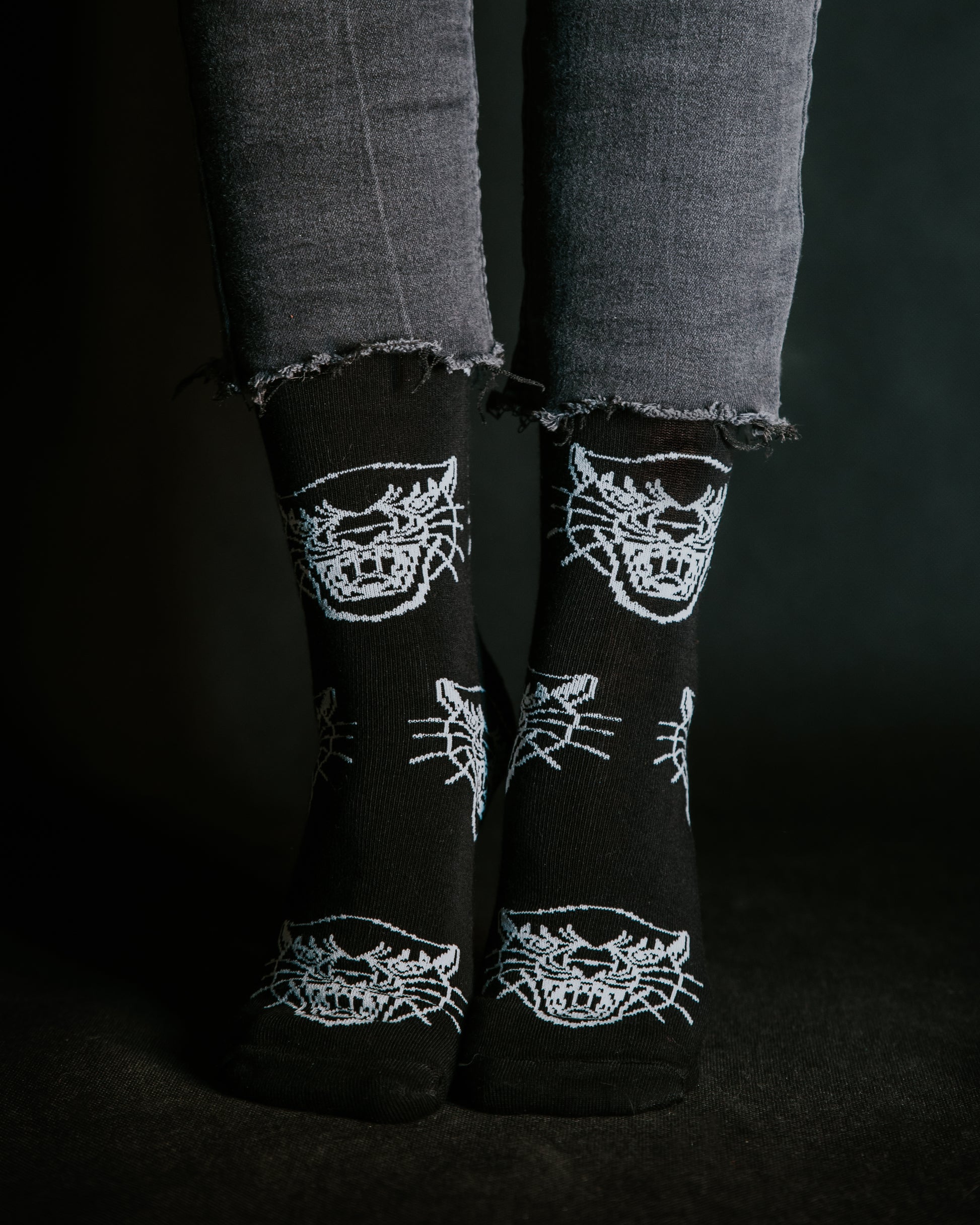 Close-up of feet wearing black LOVEINK socks with a white panther head design, paired with frayed-edge black jeans, set against a dark background.