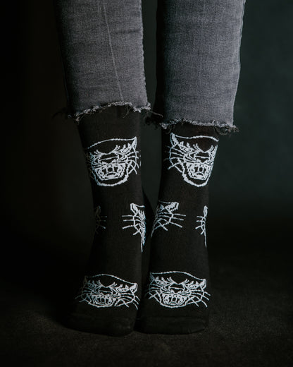 Close-up of feet wearing black LOVEINK socks with a white panther head design, paired with frayed-edge black jeans, set against a dark background.