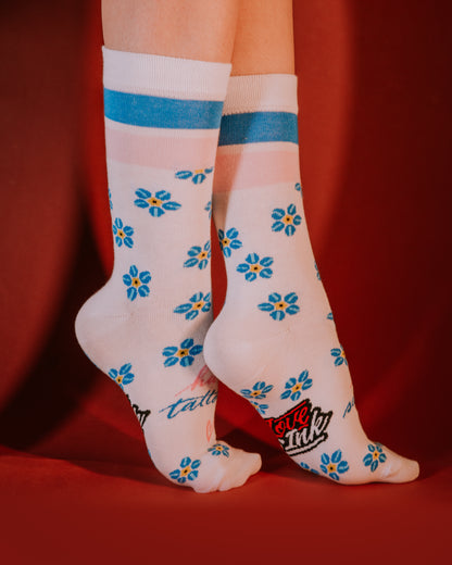Feet wearing white socks with blue and yellow flower patterns, blue and pink stripes at the top, and the Love Ink logo, posed against a red background.