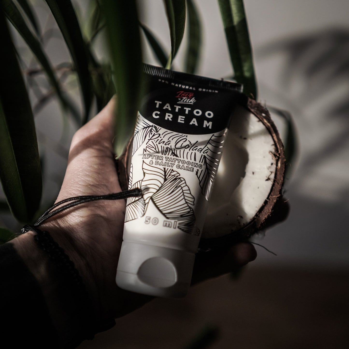 Hand holding a tube of Tattoo Cream by LoveInk with a split coconut in the background, emphasizing the product's natural ingredients.