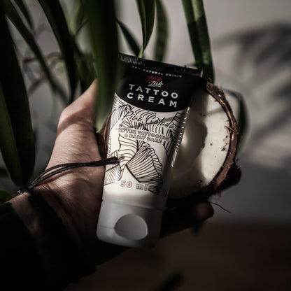 Hand holding a tube of Tattoo Cream by LoveInk with a split coconut in the background, emphasizing the product's natural ingredients.
