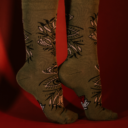 Close-up of feet wearing green LOVEINK socks featuring an intricate design with eyes and floral elements, posed against a red background.