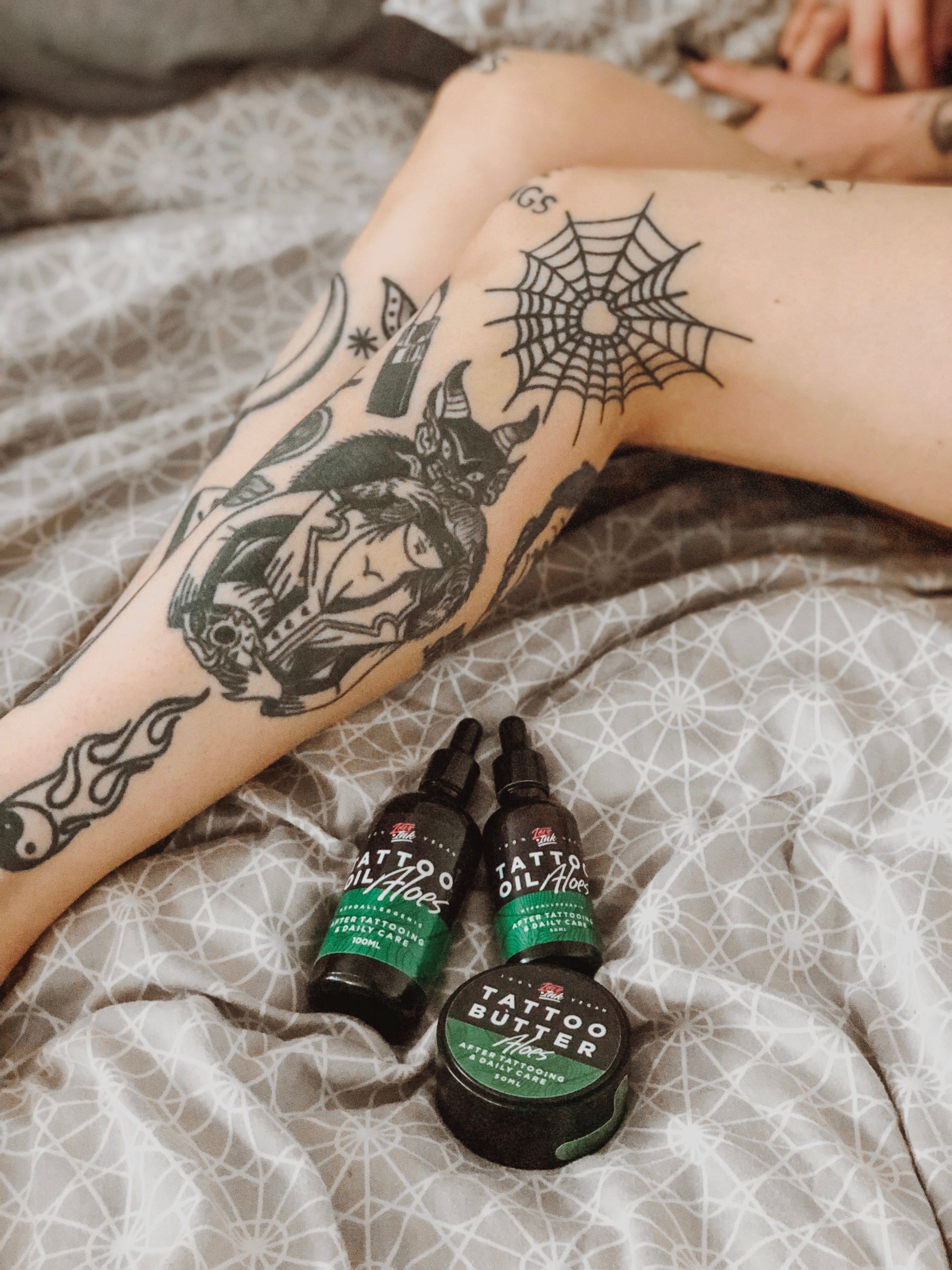 Tattooed legs on a bed with Love Ink Tattoo Butter Aloe and Tattoo Oil Aloe bottles next to them.