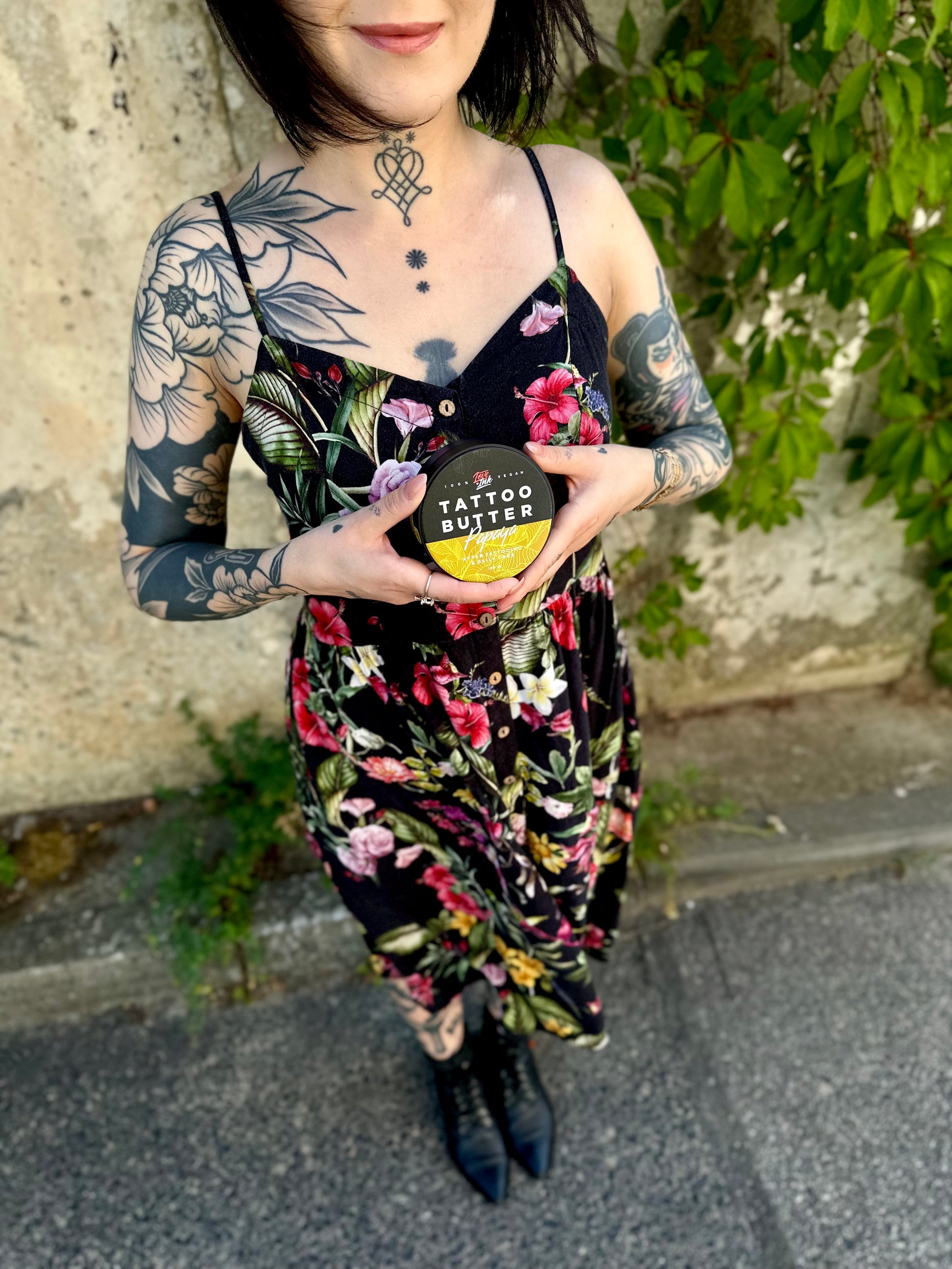 Person in a floral dress holding a jar of Tattoo Butter Papaya by LoveInk. The background features a natural setting with greenery.