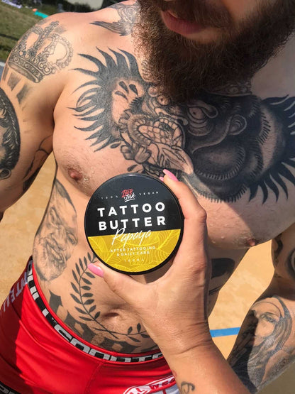 Hand holding a jar of Tattoo Butter Papaya by LoveInk against a tattooed chest. The scene highlights the product's use for tattoo care.