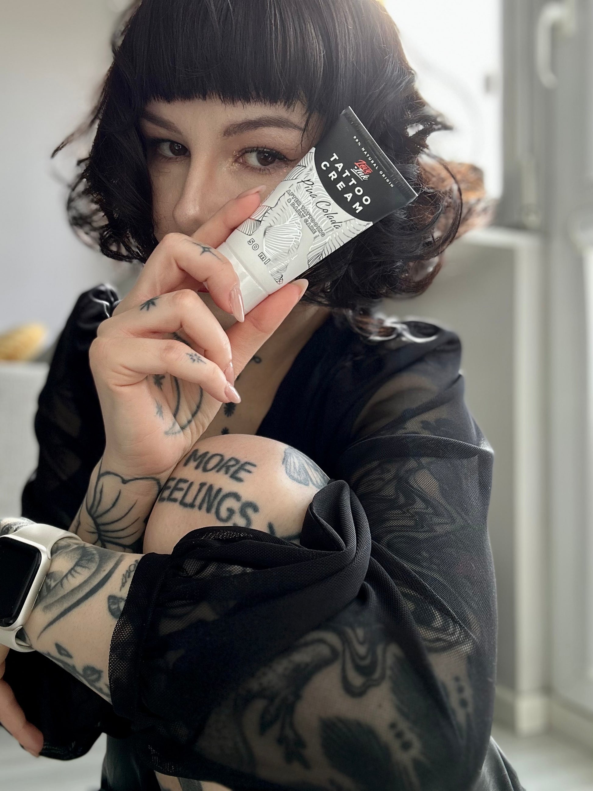 Person holding a tube of Tattoo Cream by LoveInk close to their face, showcasing detailed arm tattoos and emphasizing the product's relevance for tattoo care.