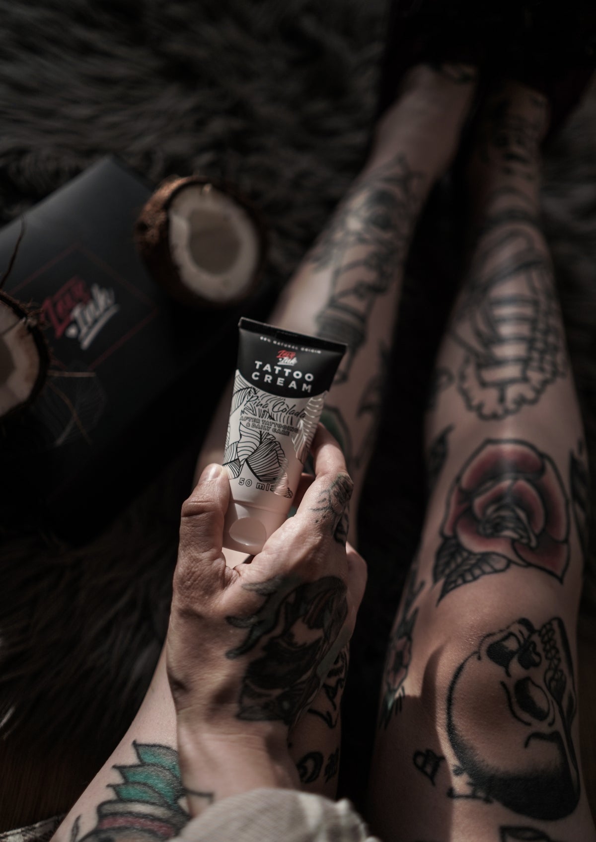 Person holding a tube of Tattoo Cream by LoveInk with detailed leg tattoos and coconut shells in the background, emphasizing the natural ingredients.