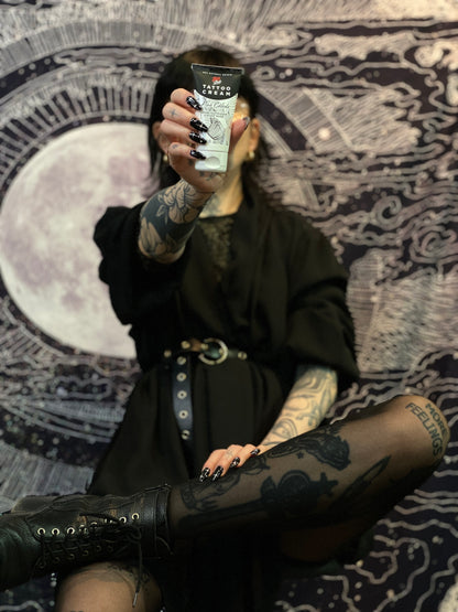 Person holding a tube of Tattoo Cream by LoveInk in front of a detailed, cosmic-themed background, highlighting the product.