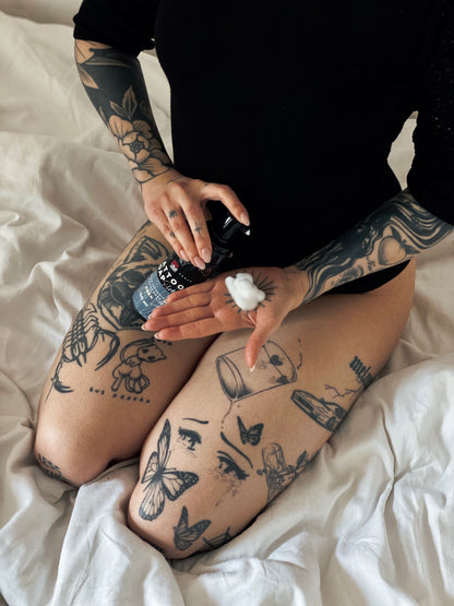 Woman in black outfit dispensing LoveInk's Tattoo Soap Silver foam onto her hand. The image shows the product being applied to the skin.